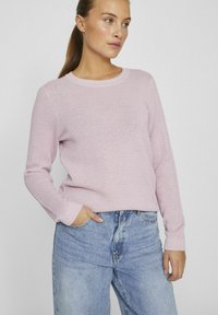 Ljust rosa stickad tröja med ett strukturerat mönster, ribbad halsringning och manschetter, tillsammans med blå denimjeans med avslappnad passform.