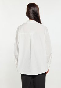 Bílé oversized košile s zadním sklady, zapínáním na knoflíky u manžet a hladkým materiálem. Spodní lem je mírně zakulacený a košile je nošena s černými kalhotami.