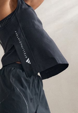 Person trägt schwarzes ärmelloses Sporttop mit "Stella McCartney"-Logo und passende schwarze Shorts vor einfarbig hellem Hintergrund.