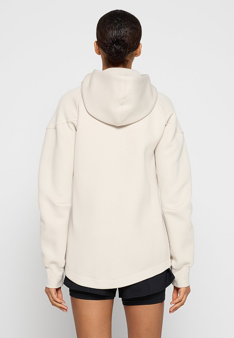 Donna con capelli scuri raccolti in un bun, che indossa una felpa oversize color crema e pantaloni shorts neri, vista di spalle su uno sfondo bianco.