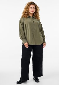 Olive green button-up shirt with a relaxed fit, long sleeves, and subtle stitching details, paired with wide black pants and black shoes.