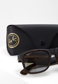Ochelari de soare Ray-Ban din plastic tortoiseshell maro închis, cu lentile polarizate, însoțiți de o carcasă texturată neagră cu un logo auriu.