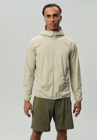 Veste légère beige clair avec capuche, fermeture éclair pleine frontale et poignets élastiques, associée à un short vert olive avec poches latérales.