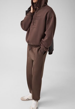 Personne portant un hoodie marron avec le texte "Los Angeles", un pantalon de survêtement marron assorti, des baskets blanches, tenant une veste sombre, debout de façon décontractée.
