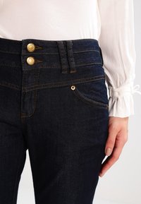 Jeans de mezclilla azul oscuro con una cintura alta, que presentan dos botones dorados, detalles de costura sutiles y una apertura de bolsillo frontal.
