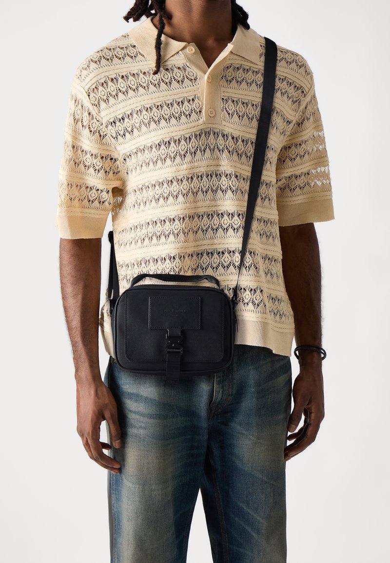 Homme portant un polo beige en dentelle à manches courtes, un jean bleu et un sac bandoulière noir avec détail boucle.