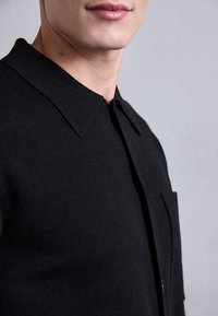 Nærbilde av en person som har på seg en svart skjorte med krage og en synlig brystlomme, mot en ensfarget bakgrunn.