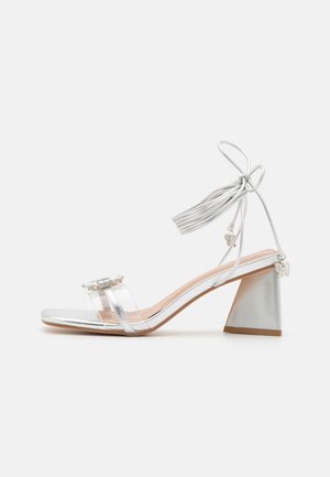 Zilveren open teen sandaal met blokhak, transparante teenband, enkelband met kwastjes en kleine vlinderbedeltjes aan de bandjes en hak.