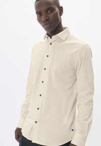 Világos bézs hosszú ujjú ing gombos gallérral, elülső gombokkal és egy kis logóval az alsó oldalon. Puha, sima szövettextúra.