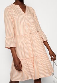 Vestido a rayas en suave melocotón y blanco, con escote en V, mangas tres cuartos y un dobladillo de volantes en capas. Tela ligera, diseño casual.