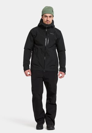 Black waterproof jacket with a zip closure and hood, paired with matching black pants. The model wears a light green beanie and black shoes.