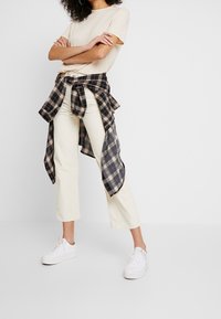 Beige t-shirt med kort ärm, en rutmönstrad skjorta i svart och vitt knuten i midjan, krämfärgade kordfläskpants och vita sneakers.