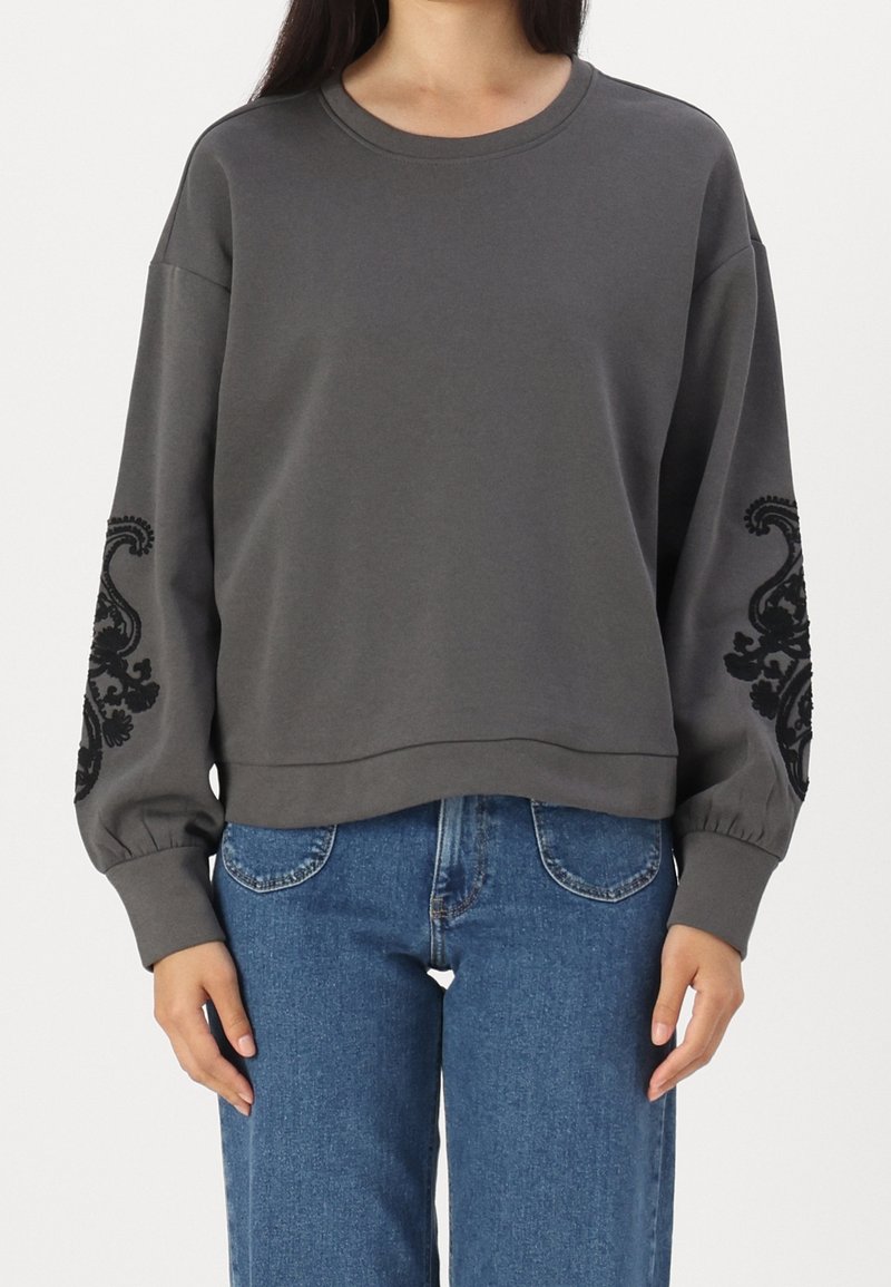 Felpa grigia con maniche lunghe e voluminose. Presenta ricami floreali neri sulle maniche, scollatura rotonda e una vestibilità rilassata. Abbinata a jeans blu.