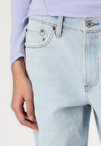 Light blue denim jeans with a relaxed fit, featuring copper rivets, contrasting stitching, and a partially visible lilac long-sleeve top.