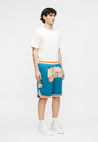 Fehér pólót, kék és narancssárga "Billionaire Boys Club" kosárlabda rövidnadrágot, fehér zoknit és fehér sportcipőt viselő férfi áll egyszerű háttér előtt.
