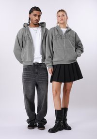 Γκρι φούτερ με φερμουάρ και κουκούλα με λογότυπο, σε συνδυασμό με μαύρο τζιν ψηλόμεσο και μαύρη πλισέ φούστα, καθώς και μαύρες μπότες μέχρι το γόνατο.