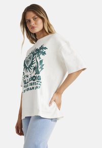 Hvid oversized t-shirt med grøn grafisk print, der viser et øje og bladdesign. Korte ærmer og en rund kant.