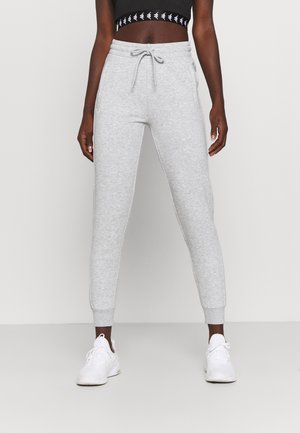 Personne portant un pantalon de survêtement gris clair avec cordon, un top court noir avec une bande blanche avec logo et des baskets blanches, debout devant un fond uni.