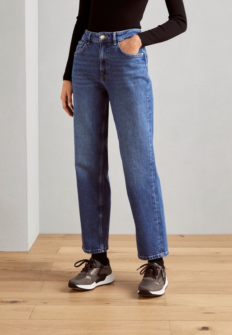 Osoba v modrých vysokých rovně střižených džínách, černém dlouhém rukávu a šedých teniskách, stojící na dřevěné podlaze.