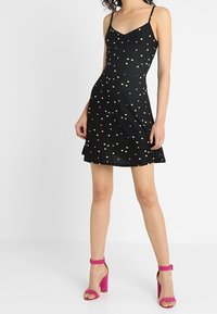 Czarna sukienka skaterka z cienkimi ramiączkami, z wielokolorowymi kropkami. Gładki materiał o sylwetce w kształcie litery A. Połączona z różowymi sandałami na blokowym obcasie.