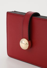 Punane nahast rahakott, millel on sile tekstuur, kuldne ümmargune sulgur, millel on kiri "Furla", ja kontrastsete mustade õmblustega ääred.