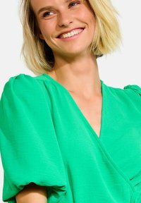 Zelené zavinovacie šaty s puff rukávmi, vyrobené z ľahkého materiálu. Majú hlboký V-výstrih a hladkú textúru. Svetlá farba zvyšuje viditeľnosť.
