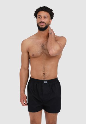 Schwarze Boxershorts aus einem leichten Stoff mit einem elastischen Bund, versehen mit einem Logo auf der Vorderseite. Das Modell steht oberkörperfrei vor einem neutralen Hintergrund.