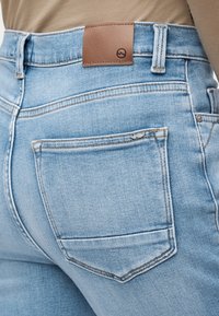 Κοντινό πλάνο τσέπης πίσω και ζώνης από ανοιχτό γαλάζιο τζιν με ταμπά δερμάτινη ετικέτα μάρκας, φορεμένα με μπεζ τοπ.