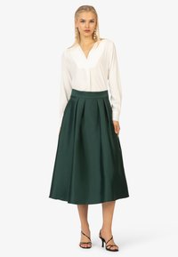 Witte blouse met v-hals, gecombineerd met een groene, geplooide midi-jurk. Zwarte sandalen met bandjes maken de look compleet. Glad stof en strakke lijnen.