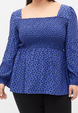 Kobieta ubrana w niebieską bluzkę w czarne kropki, z długimi bufkami i ściąganym kwadratowym dekoltem, w zestawieniu z czarnymi spodniami.