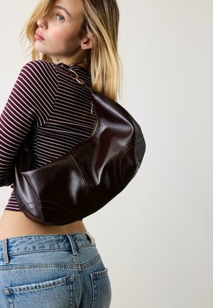 Mlada ženska s svetlimi lasmi, oblečena v črtasto grozdno rdečo majico in modre kavbojke, nosi veliko temno rjavo usnjeno torbo čez ramo.