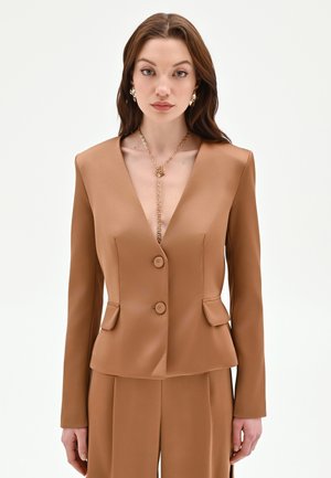 Femme portant un blazer marron sur mesure à col en V profond, un pantalon assorti, un collier chaîne en or et des boucles d'oreilles en or sur un fond uni.