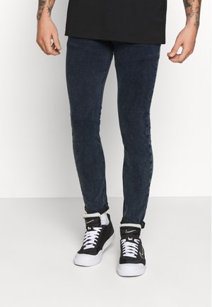Person trägt dunkelblaue Skinny-Jeans und schwarze High-Top-Sneaker mit weißen Schnürsenkeln und Sohlen, steht vor einem einfarbigen Hintergrund.