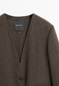 Brun uldfrakke med V-halsdesign, enkelt sort knaplukning og en blød, struktureret finish. Mærke synligt indeni halsudskæringen.