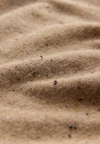 Ingrandimento di un tessuto marrone chiaro con piccole macchie scure e un motivo a maglia testurizzato che mostra pieghe e ombre sottili.