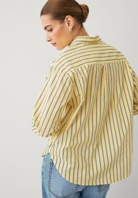 Chemise rayée jaune avec des rayures horizontales bleues, col boutonné et coupe décontractée. Comprend un ourlet arrondi et une texture douce.