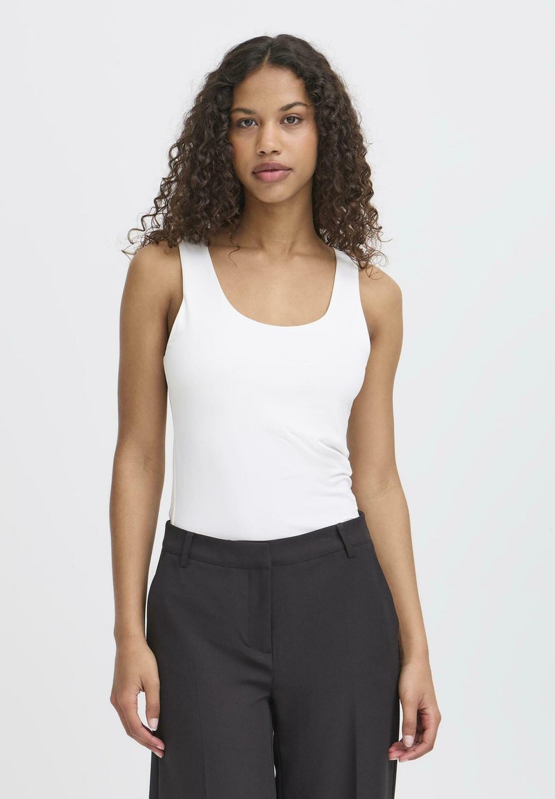 Femme aux cheveux bouclés portant un haut blanc sans manches et un pantalon noir, debout devant un fond clair uni.