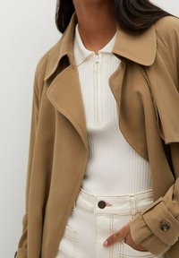 Helle beige Trenchcoat mit weicher Textur, übergroßem Kragen und Knopfmanschetten, getragen über einem weißen gerippten Top und cremfarbenen Hosen.