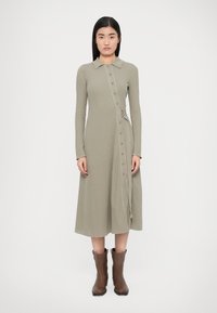 Lang ribstrikket kjole i afdæmpet grøn med krave, ærmer i fuld længde, knaplukning foran og bælte som detalje. Kombineret med brune ankelstøvler.