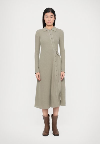 Robe longue côtelée en vert doux, avec col, manches longues, boutonnage devant et ceinture. Associée à des bottines marron.