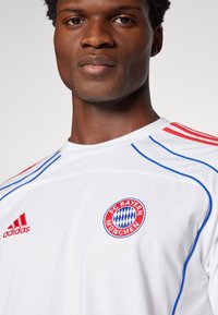Biała koszulka z krótkim rękawem z czerwonymi i niebieskimi akcentami. Posiada logo FC Bayern München oraz branding Adidas na lewej piersi i ramieniu.
