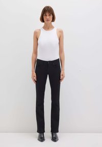 Donna in piedi contro un muro bianco che indossa un top bianco senza maniche, pantaloni neri a gamba dritta e stivaletti neri.
