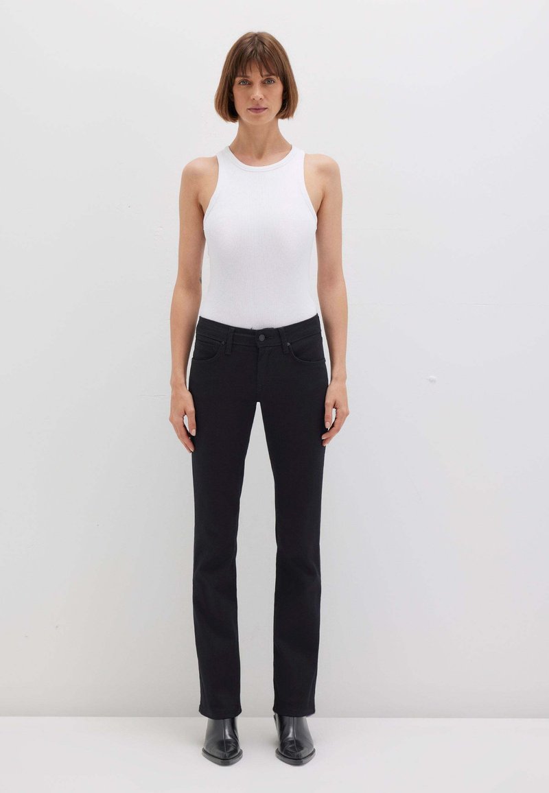 Donna in piedi contro un muro bianco che indossa un top bianco senza maniche, pantaloni neri a gamba dritta e stivaletti neri.