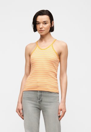 Jeune femme aux cheveux courts et foncés portant un haut sans manches rayé jaune et orange et un jean gris clair, debout devant un fond blanc.