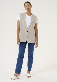 Beige sleeveless vest with a single black button, worn over a white T-shirt, paired with blue jeans and white heeled sandals.