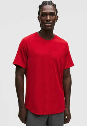 Homme aux cheveux tressés portant un t-shirt rouge uni à manches courtes et un pantalon gris, debout devant un fond blanc.