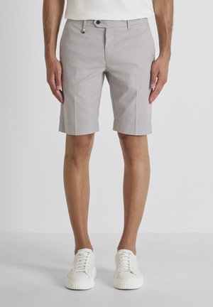 Hombre con pantalones cortos a medida de color gris claro y zapatillas blancas, de pie frente a un fondo blanco liso.