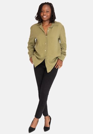 Glunderende vrouw met gevlochten haar, gekleed in een olijfgroene blouse met knopen, zwarte skinny jeans en zwarte puntige hakken, staand met gekruiste benen.