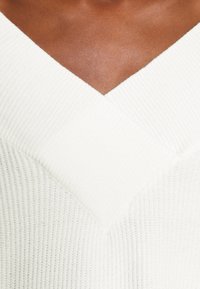 Vit ribbad stickad tröja med djup V-ringning och strukturerad design, som visar mjukt material med en åtsittande silhuett.