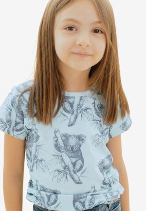 Junges Mädchen mit glatten braunen Haaren, das ein hellblaues T-Shirt mit Mustern von Skizzen von Koalas auf Baumzweigen trägt.